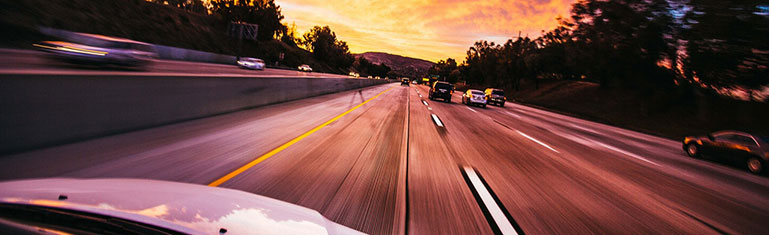 Cars speeding down the highway with a sunset on the horizon.
